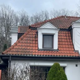 Czerwona dachówka karpiówka na białym domu z oknem mansardowym, widok na fragment elewacji i drzewa bez liści w tle.