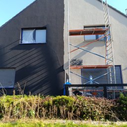 Darfloor - Dwukolorowa elewacja domu jednorodzinnego w trakcie prac. Część ściany pomalowana na grafitowo, druga połowa na jasny odcień. Okna zabezpieczone folią. Przy ścianie ustawione rusztowanie z butelką...