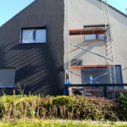 Dwukolorowa elewacja domu jednorodzinnego w trakcie prac. Część ściany pomalowana na grafitowo, druga połowa na jasny odcień. Okna zabezpieczone folią. Przy ścianie ustawione rusztowanie z butelką...