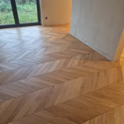 Ułożona jodełka francuska z drewnianych desek podłogowych w jasnym odcieniu dębu, widoczne fragmenty niezamalowanej ściany i duże okno z widokiem na zieleń.