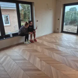 Dwie kobiety siedzą na parapecie okna, podziwiając nowo ułożony parkiet w jodełkę w jasnym, przestronnym pokoju z dużymi oknami i surowymi ścianami.