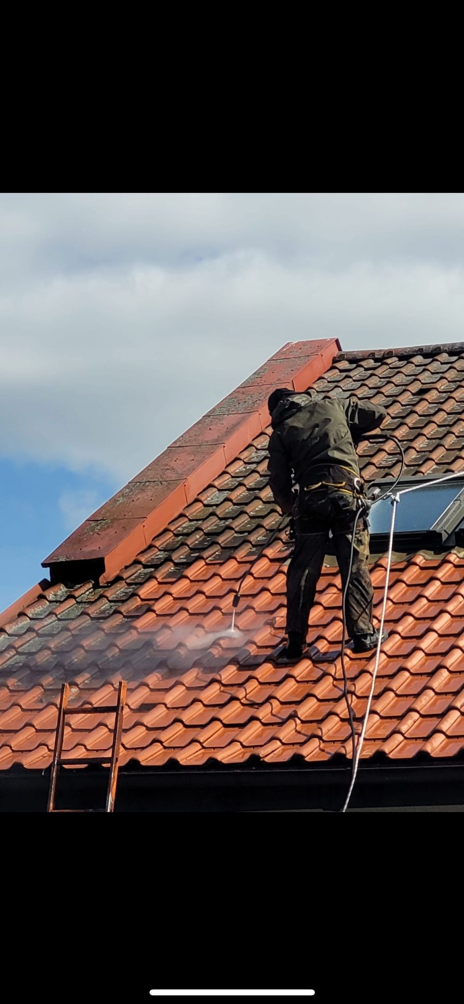 Pracownik w uprzęży, z wysokociśnieniową myjką, czyści czerwoną dachówkę na stromym dachu, z widocznym oknem dachowym i fragmentem drabiny.