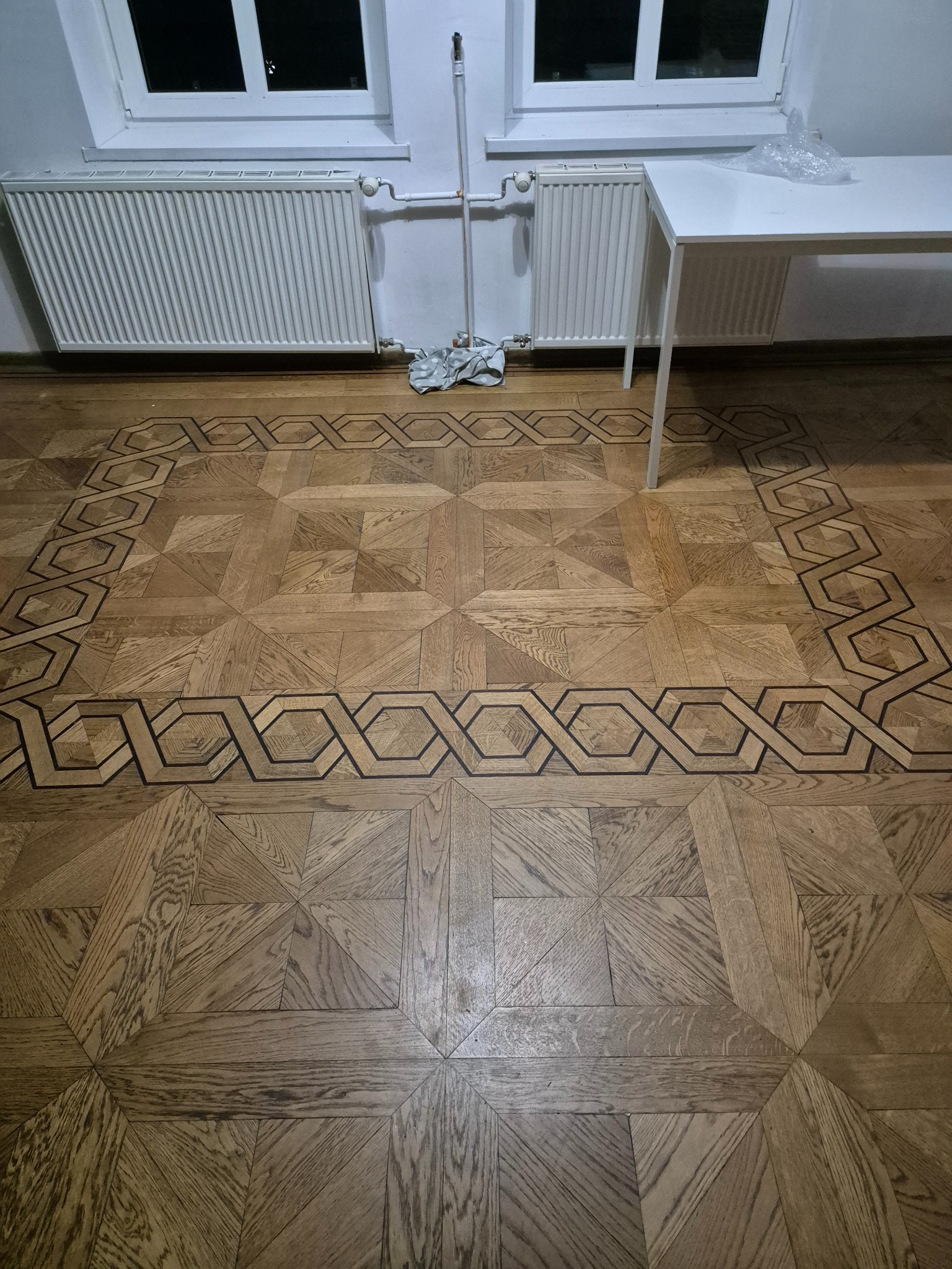 Ułożona parkietowa podłoga z geometrycznym wzorem, widoczne grzejniki i fragment białego stołu w tle.