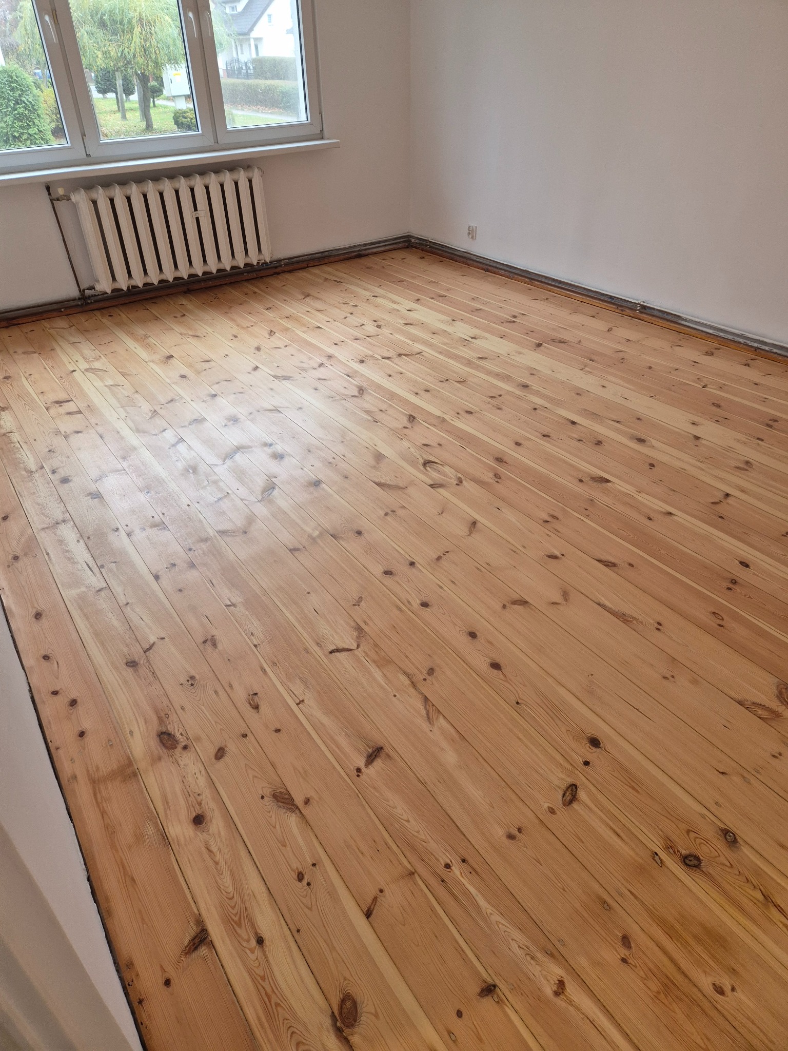 Jasna, odnowiona podłoga z desek sosnowych w pustym pokoju z białymi ścianami i grzejnikiem żeberkowym pod oknem.