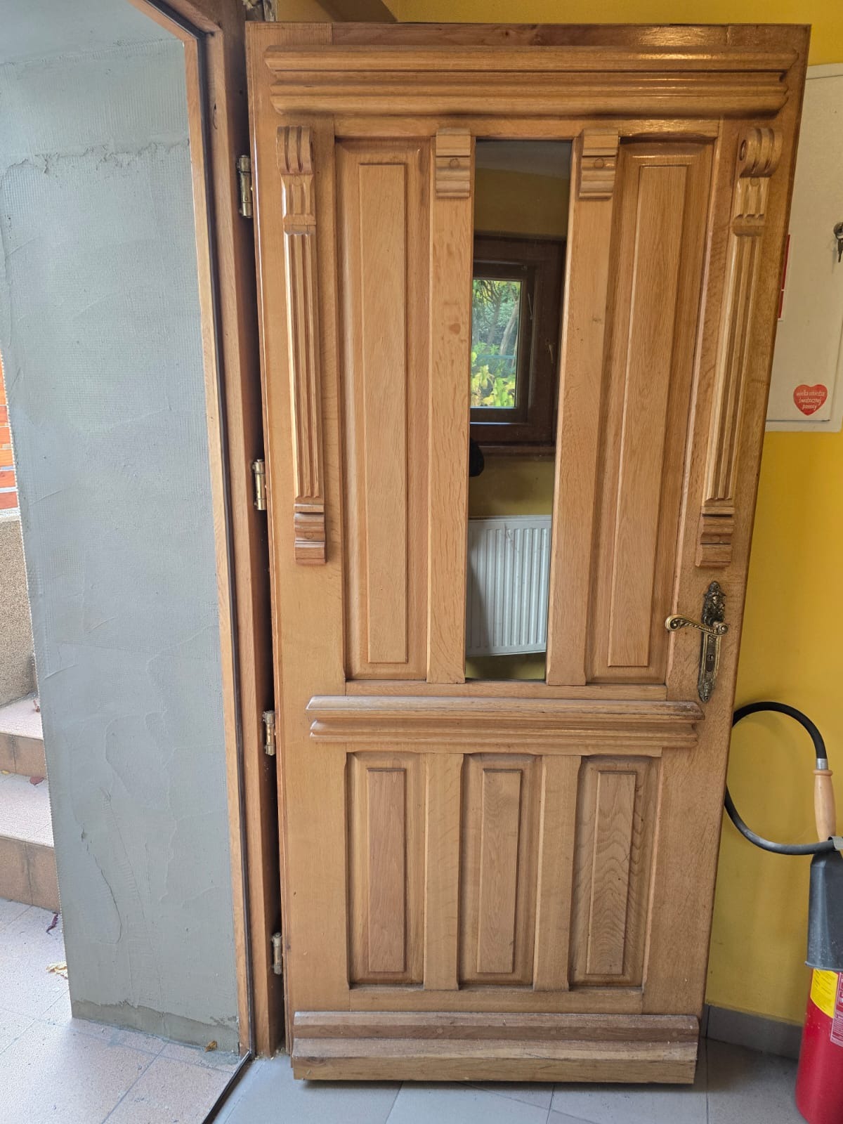 Otwarta, drewniana, ozdobna brama garażowa z widocznym wnętrzem budynku i grzejnikiem; po prawej stronie gaśnica.