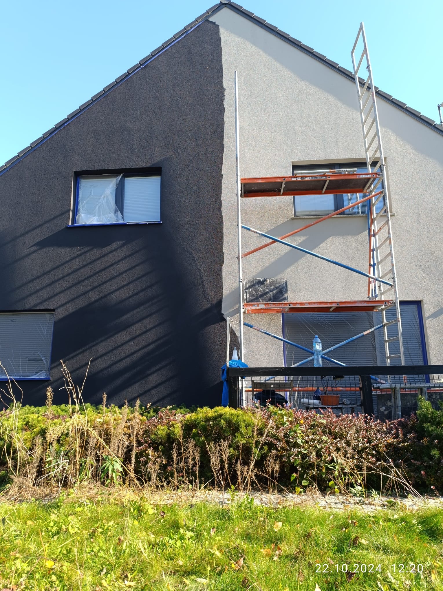 Dwukolorowa elewacja domu jednorodzinnego w trakcie prac. Część ściany pomalowana na grafitowo, druga połowa na jasny odcień. Okna zabezpieczone folią. Przy ścianie ustawione rusztowanie z butelką...