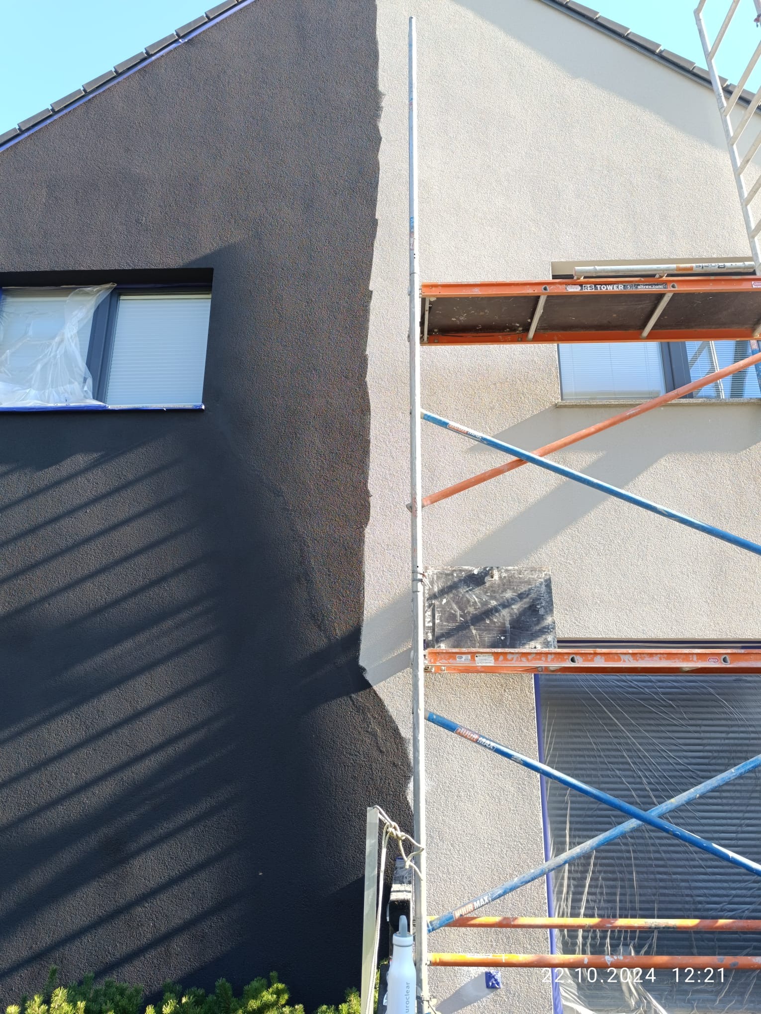 Elewacja domu w trakcie malowania, widoczne rusztowanie, okno zabezpieczone folią, część ściany w kolorze grafitowym, druga część w jasnym odcieniu.