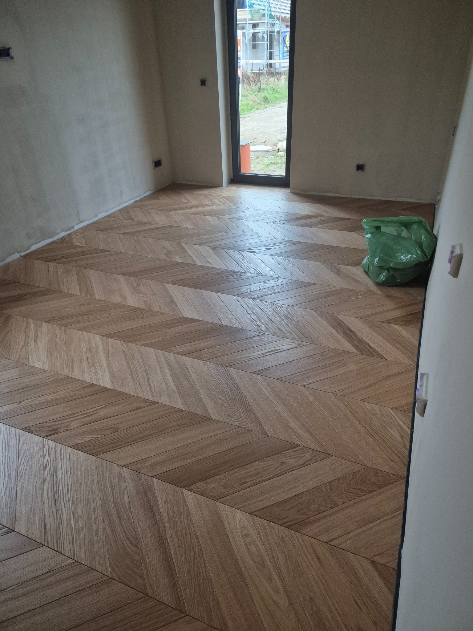 Ułożona w jodełkę, jasnobrązowa drewniana podłoga w nowym pokoju, z widocznym workiem na śmieci i surowymi ścianami.