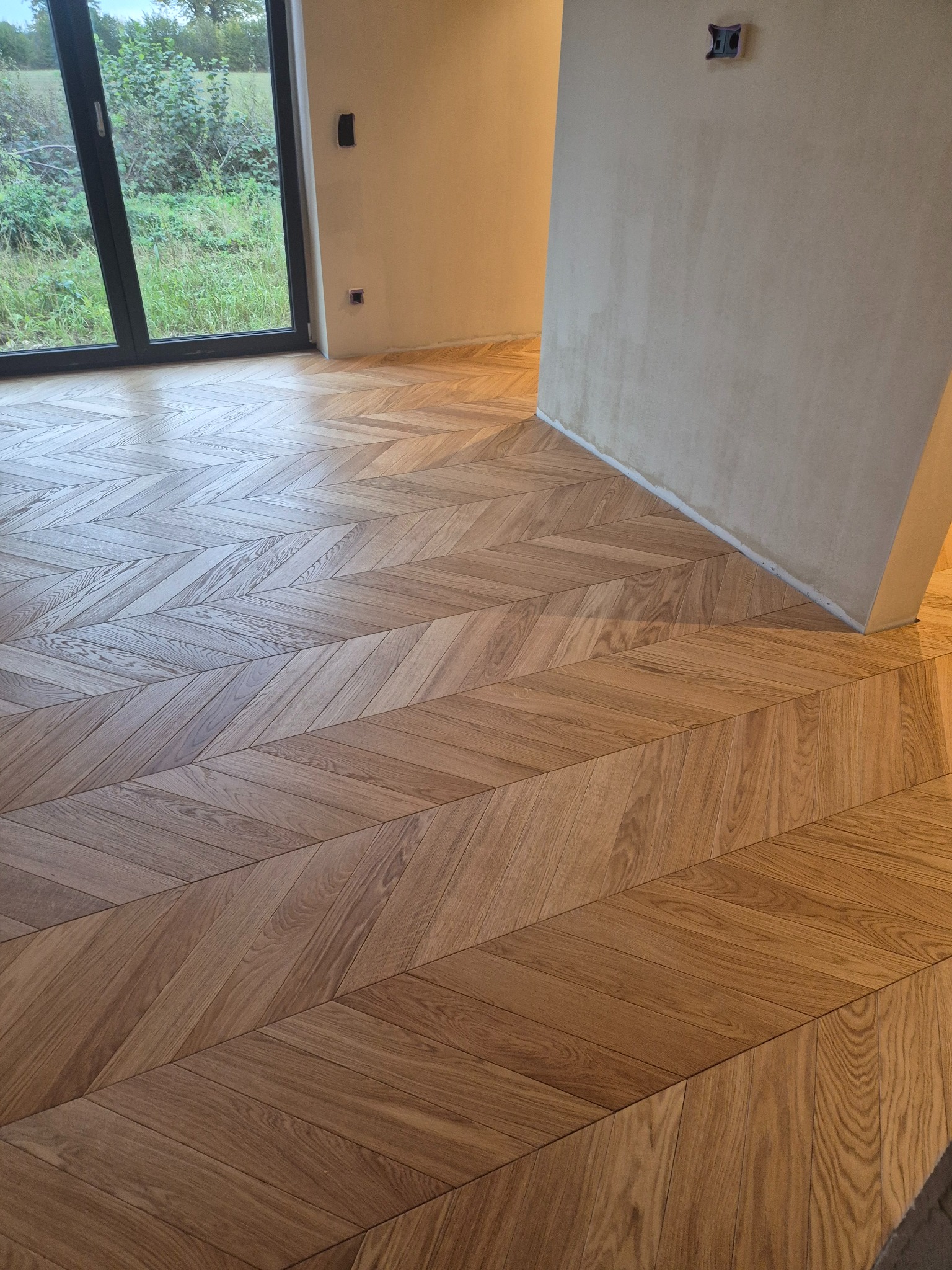 Ułożona jodełka francuska z drewnianych desek podłogowych w jasnym odcieniu dębu, widoczne fragmenty niezamalowanej ściany i duże okno z widokiem na zieleń.