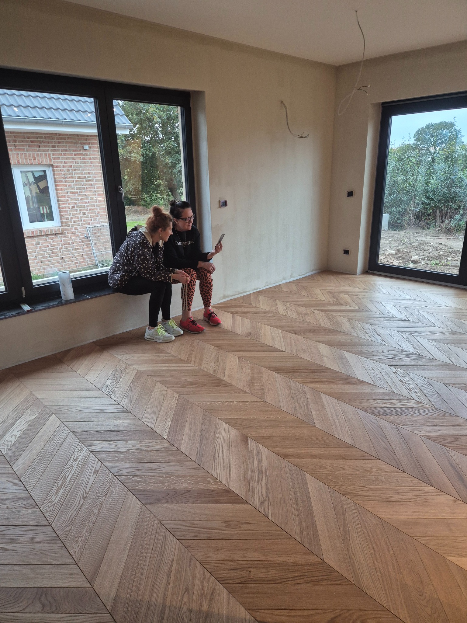 Dwie kobiety siedzą na parapecie okna, podziwiając nowo ułożony parkiet w jodełkę w jasnym, przestronnym pokoju z dużymi oknami i surowymi ścianami.