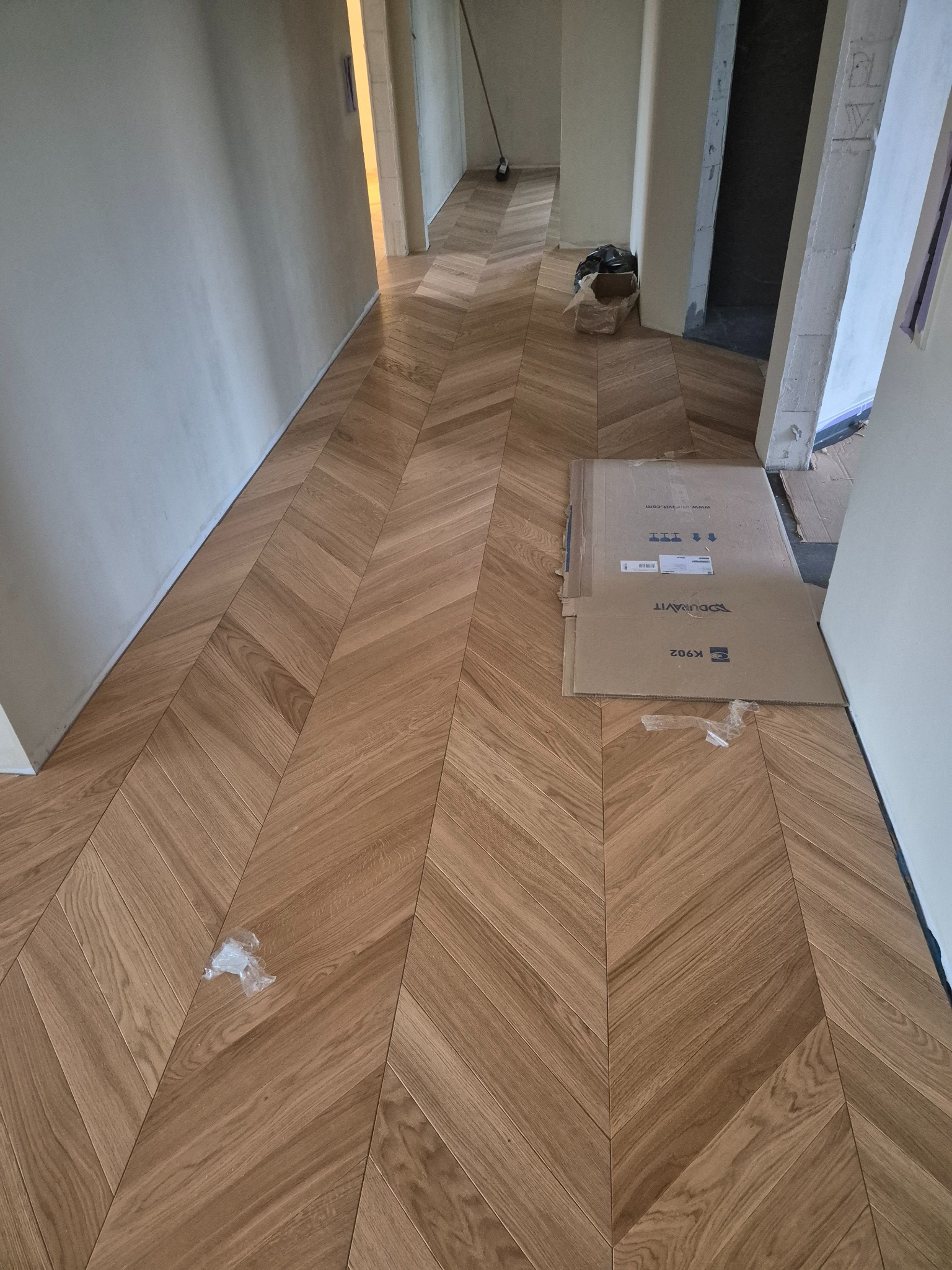 Ułożony parkiet w jodełkę, widoczne kartony po produkcie Duravit K902 i drobne śmieci budowlane w korytarzu podczas remontu.