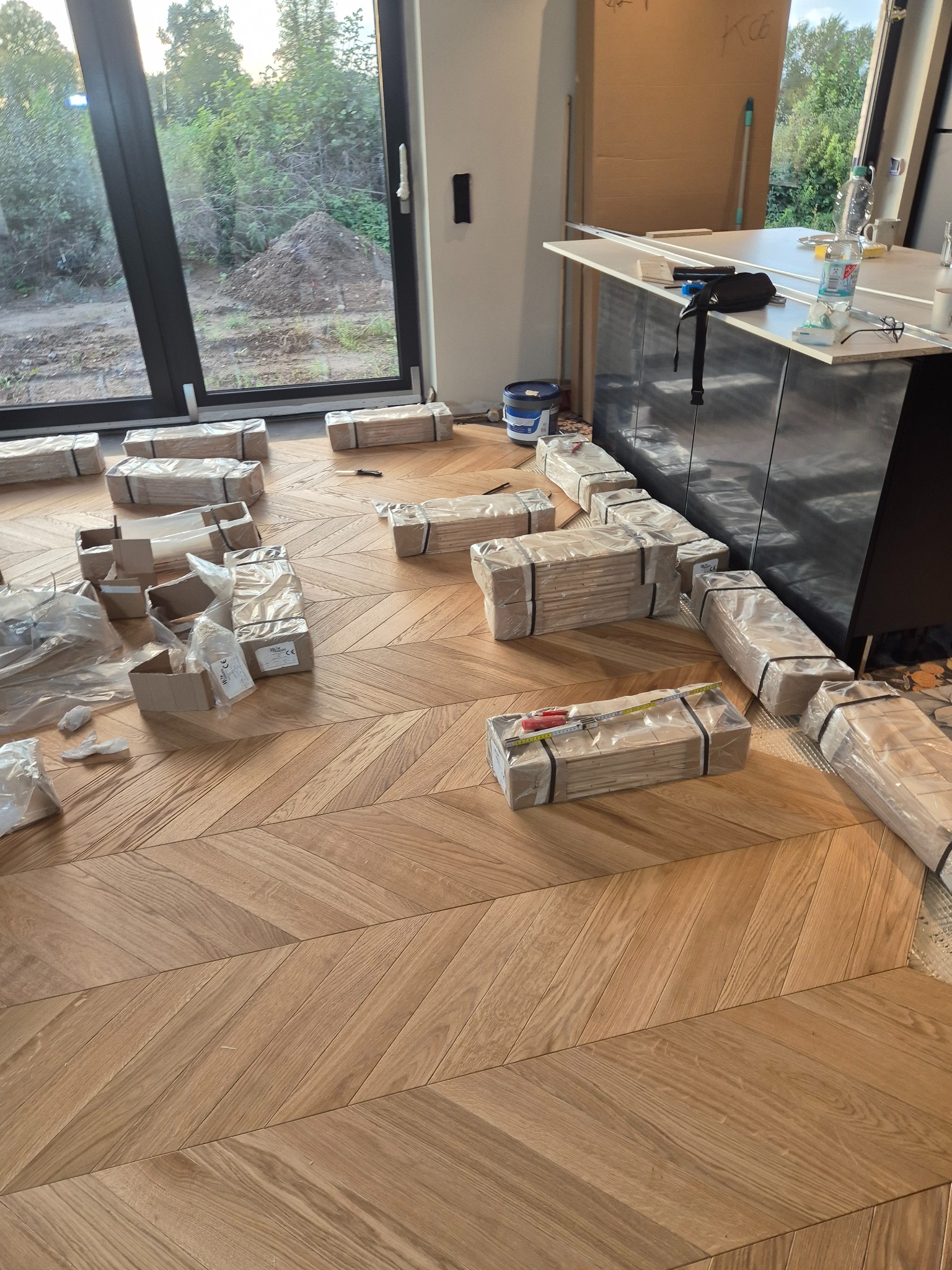 Ułożony parkiet w jodełkę z paczkami niezamontowanych desek, narzędzia pomiarowe i wiadro farby w tle.