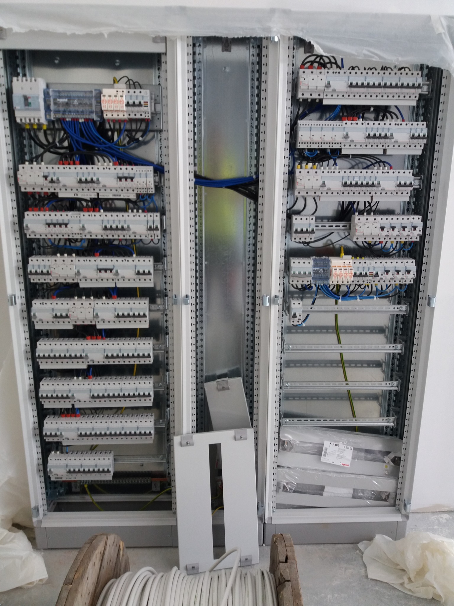 Rozdzielnica elektryczna w trakcie montażu, widoczne kable, zabezpieczenia i elementy konstrukcyjne. Na dole szpula z przewodem. Częściowo zamontowane panele.