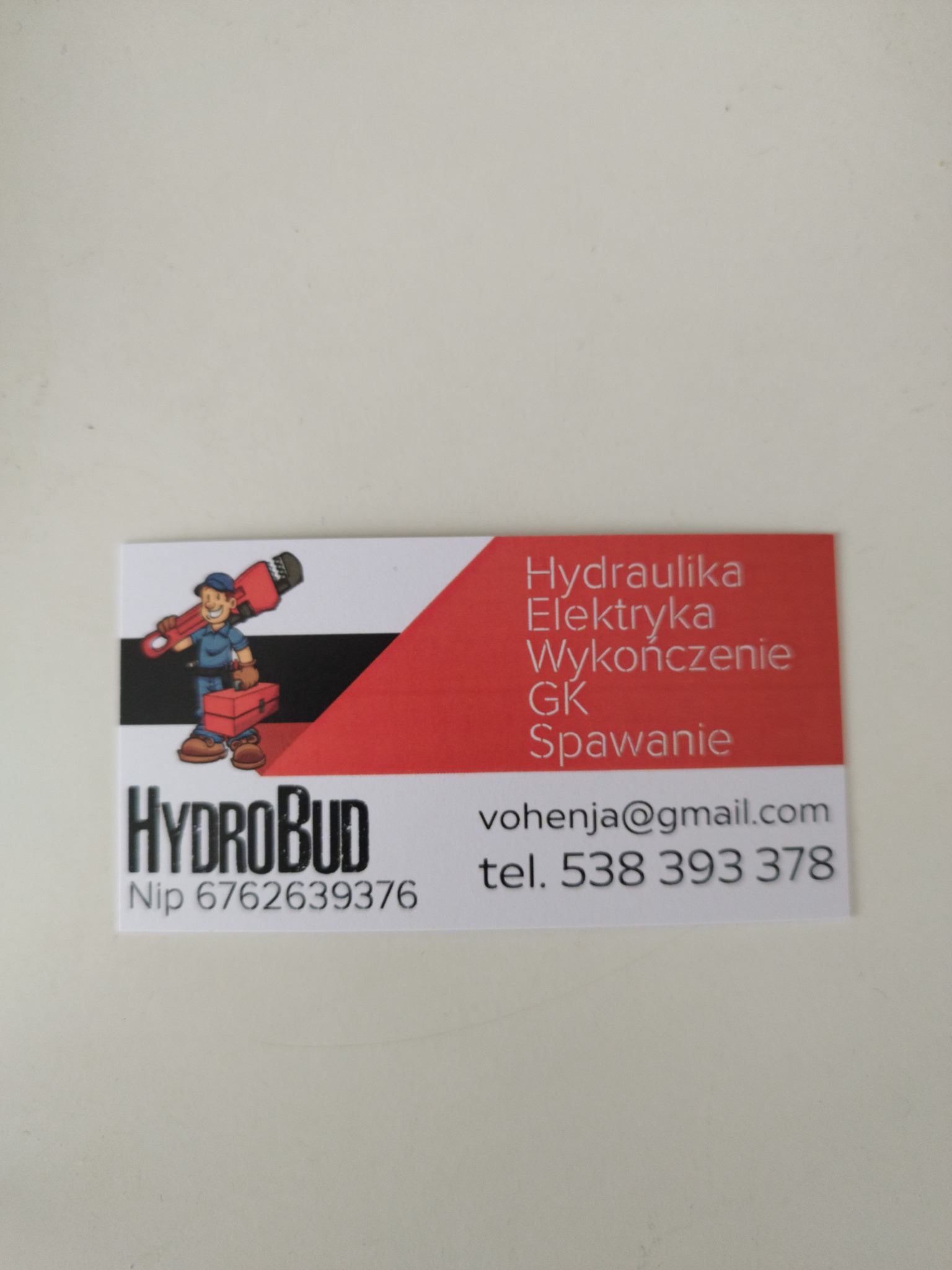 Wizytówka firmy HydroBud z ilustracją hydraulika z kluczem i skrzynką narzędziową, oferującej usługi hydrauliczne, elektryczne, wykończeniowe, GK i spawalnicze, z podanym adresem e-mail i numerem...