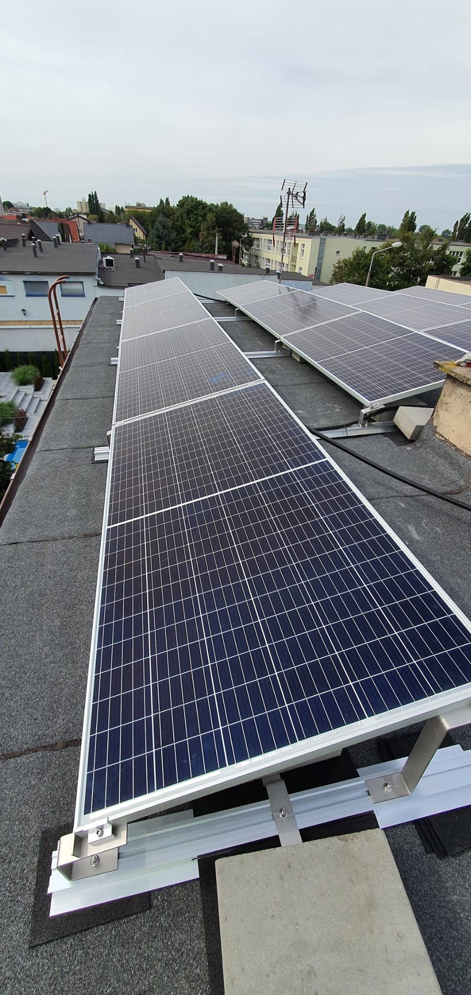 Panele słoneczne zamontowane na płaskim dachu pokrytym papą, widoczne srebrne profile montażowe i betonowe obciążniki, w tle zabudowa miejska i pochmurne niebo.