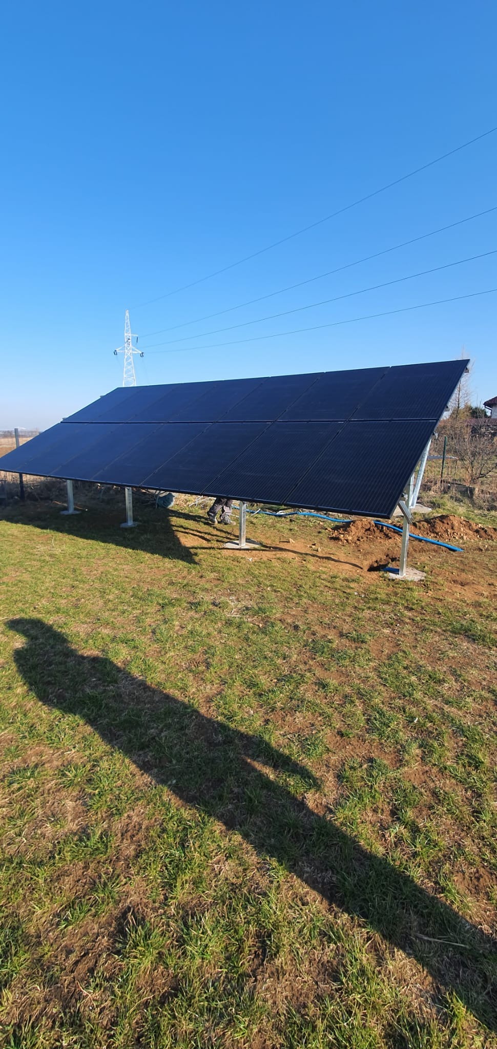 Nisko osadzony panel słoneczny na metalowych wspornikach, częściowo zasłonięty cieniem, z widocznymi kablami i wykopaną ziemią wokół podstawy, na tle błękitnego nieba i linii wysokiego napięcia.