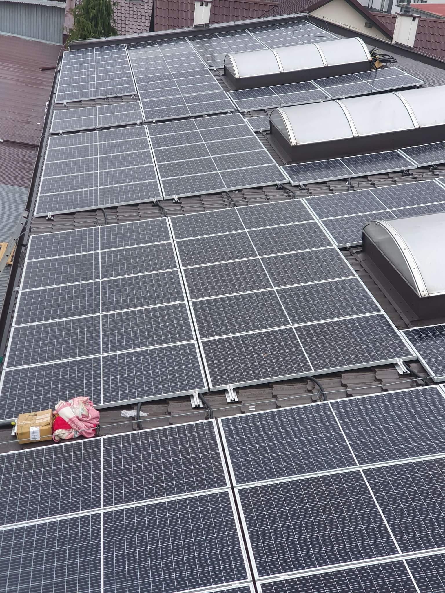 Panele słoneczne na dachu z brązową dachówką, widoczne okna dachowe i karton z kocem na panelach, instalacja w trakcie.