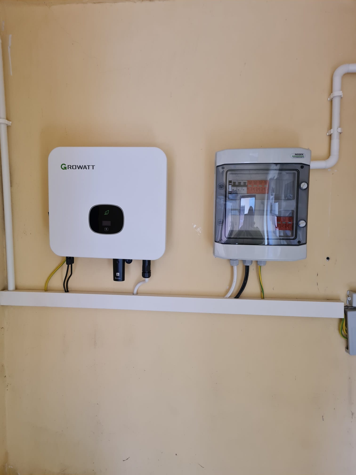 Zamontowany biały falownik marki Growatt z podłączonymi przewodami i szara skrzynka bezpiecznikowa Noark na kremowej ścianie, instalacja na białej listwie przypodłogowej.