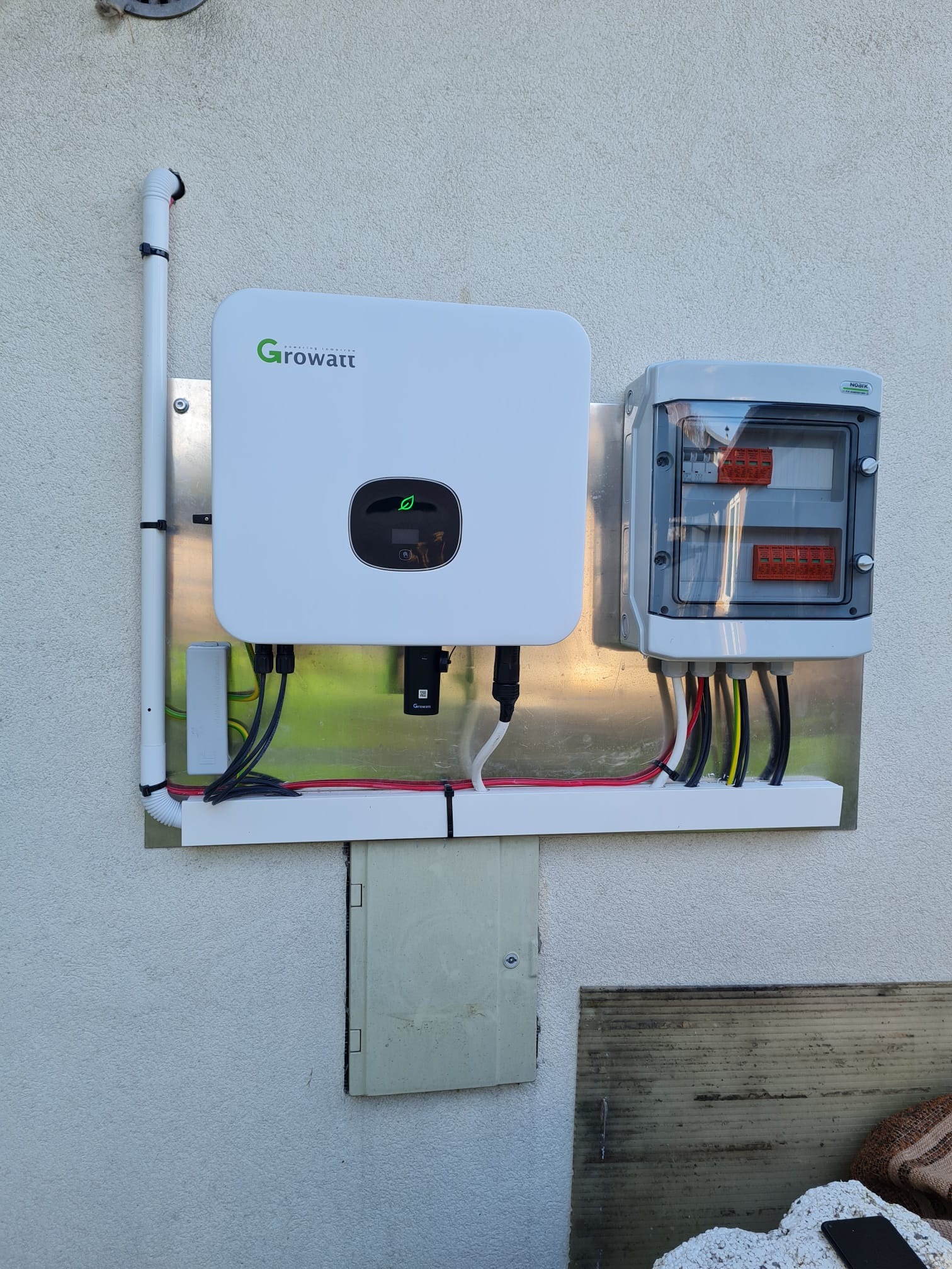 Biały falownik Growatt zamontowany na metalowej płycie obok skrzynki elektrycznej z widocznymi przewodami na elewacji budynku.