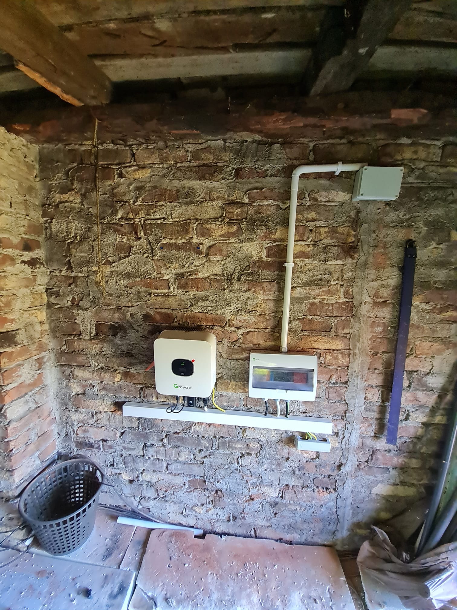 Wnętrze z surową ceglaną ścianą, na której zamontowano biały inwerter Growatt, skrzynkę rozdzielczą i białe rury instalacyjne. Widoczne drewniane belki stropowe i fragment kosza.