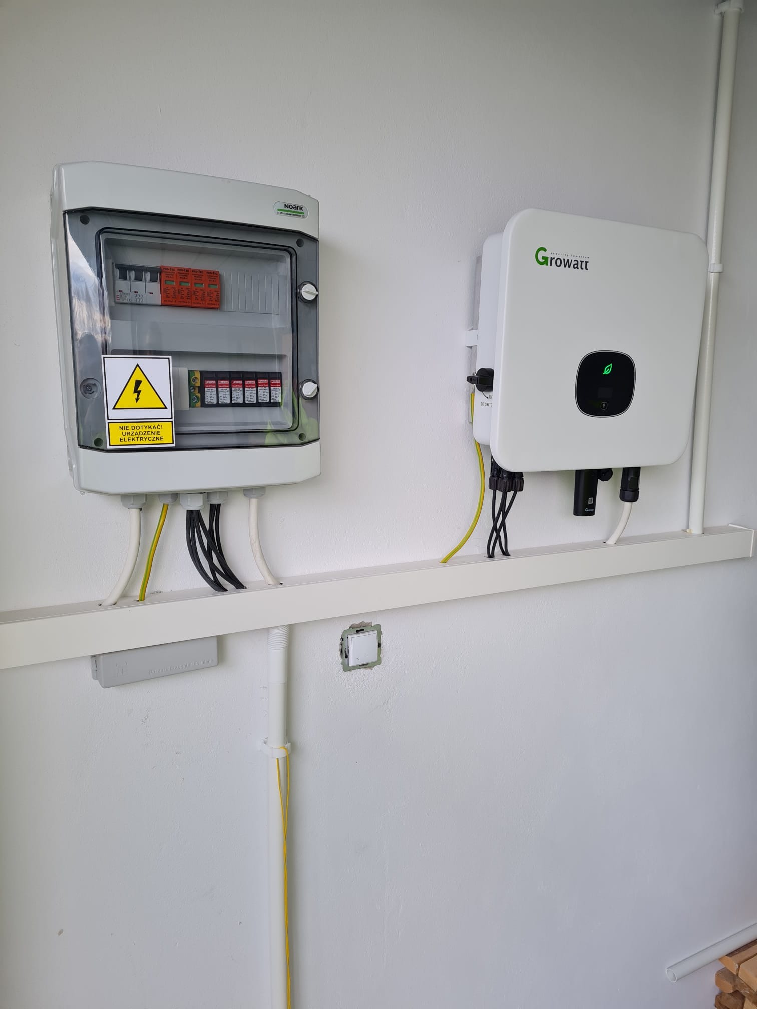 Biała ściana z zamontowaną szarą skrzynką elektryczną Noark z żółtym znakiem ostrzegawczym i bezpiecznikami oraz białym falownikiem Growatt z zielonym logo liścia, połączone przewodami na białej...
