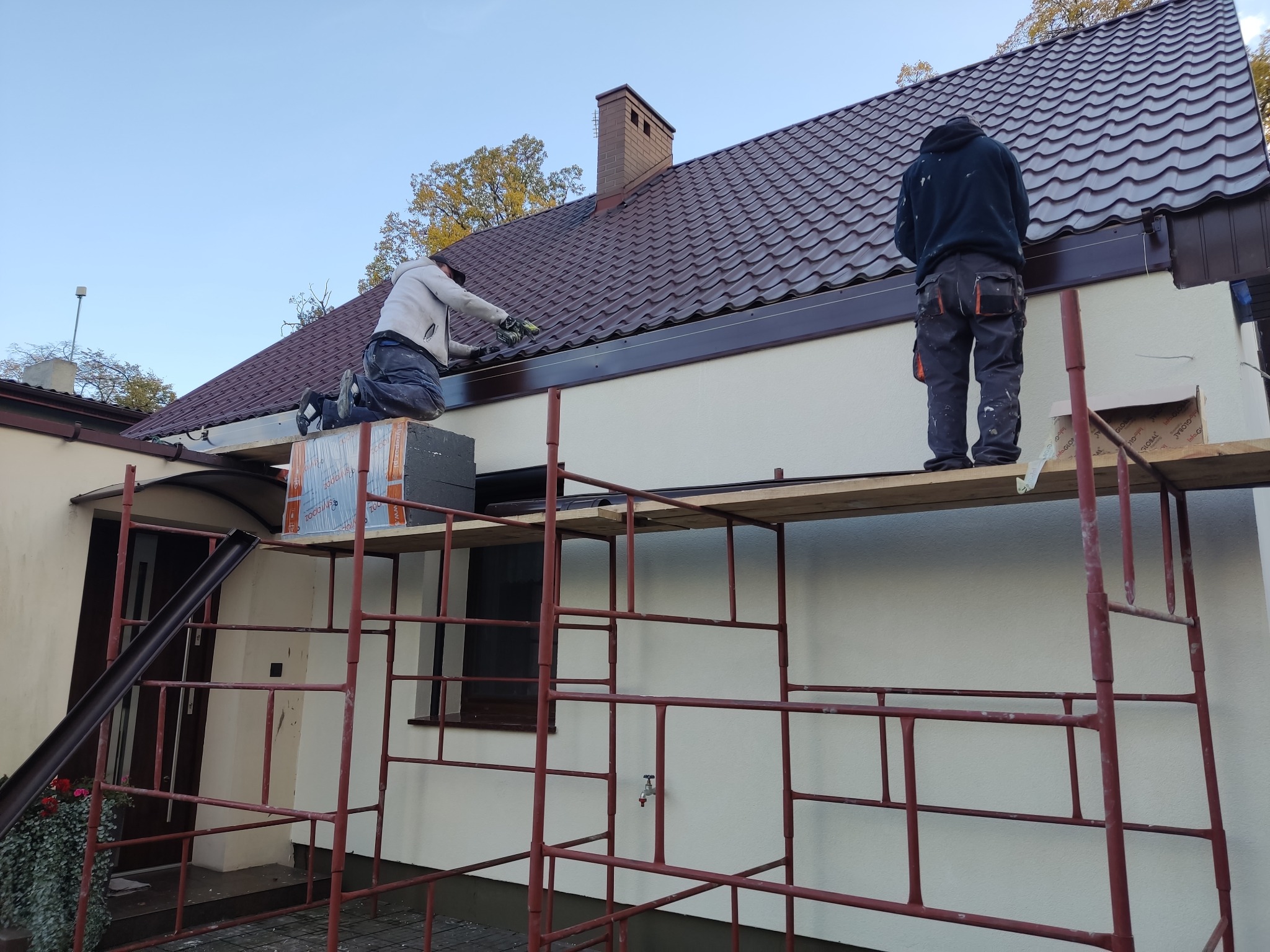 Prace malarskie elewacji domu jednorodzinnego z rusztowania, widoczne dwie osoby podczas malowania ciemnobrązowych elementów wykończeniowych dachu i elewacji.