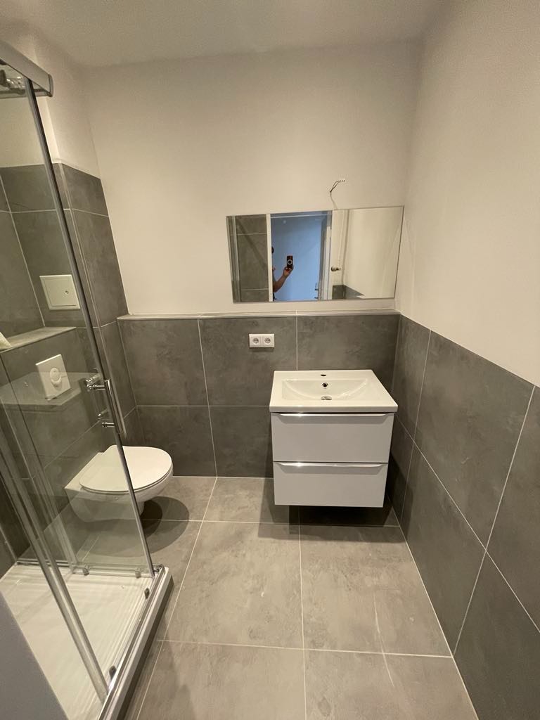 Nowoczesna łazienka z szarą glazurą, wisząca toaleta i umywalka z szafką, widok na lustro z odbiciem osoby robiącej zdjęcie.