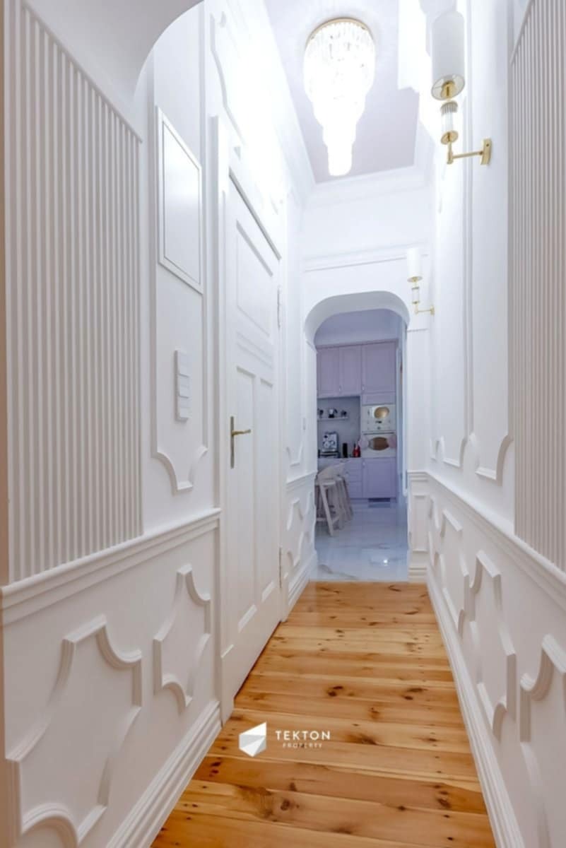 Jasny korytarz z drewnianą podłogą i białymi ścianami z dekoracyjnymi panelami, widok na fioletową kuchnię przez łuk.