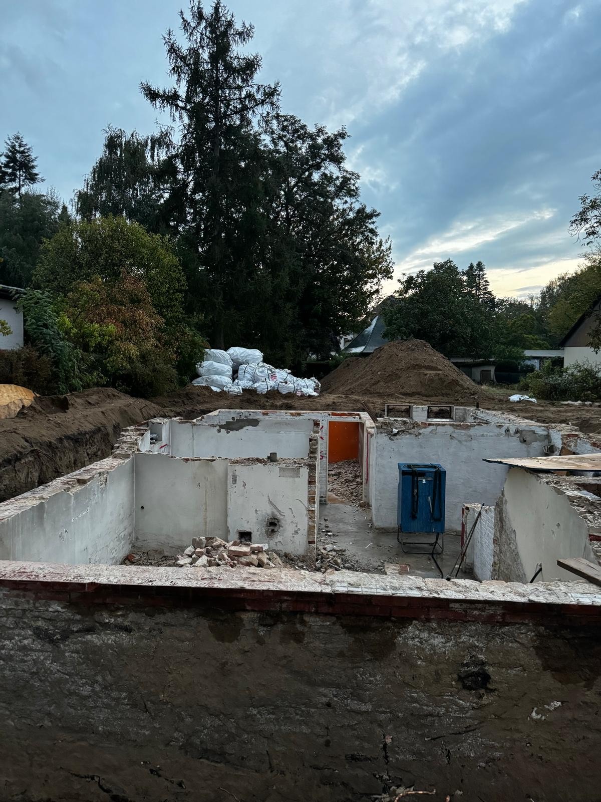 Fundamenty po rozbiórce budynku, widoczne fragmenty ścian, gruz, worki z materiałem budowlanym i pryzma ziemi w tle, pochmurne niebo.