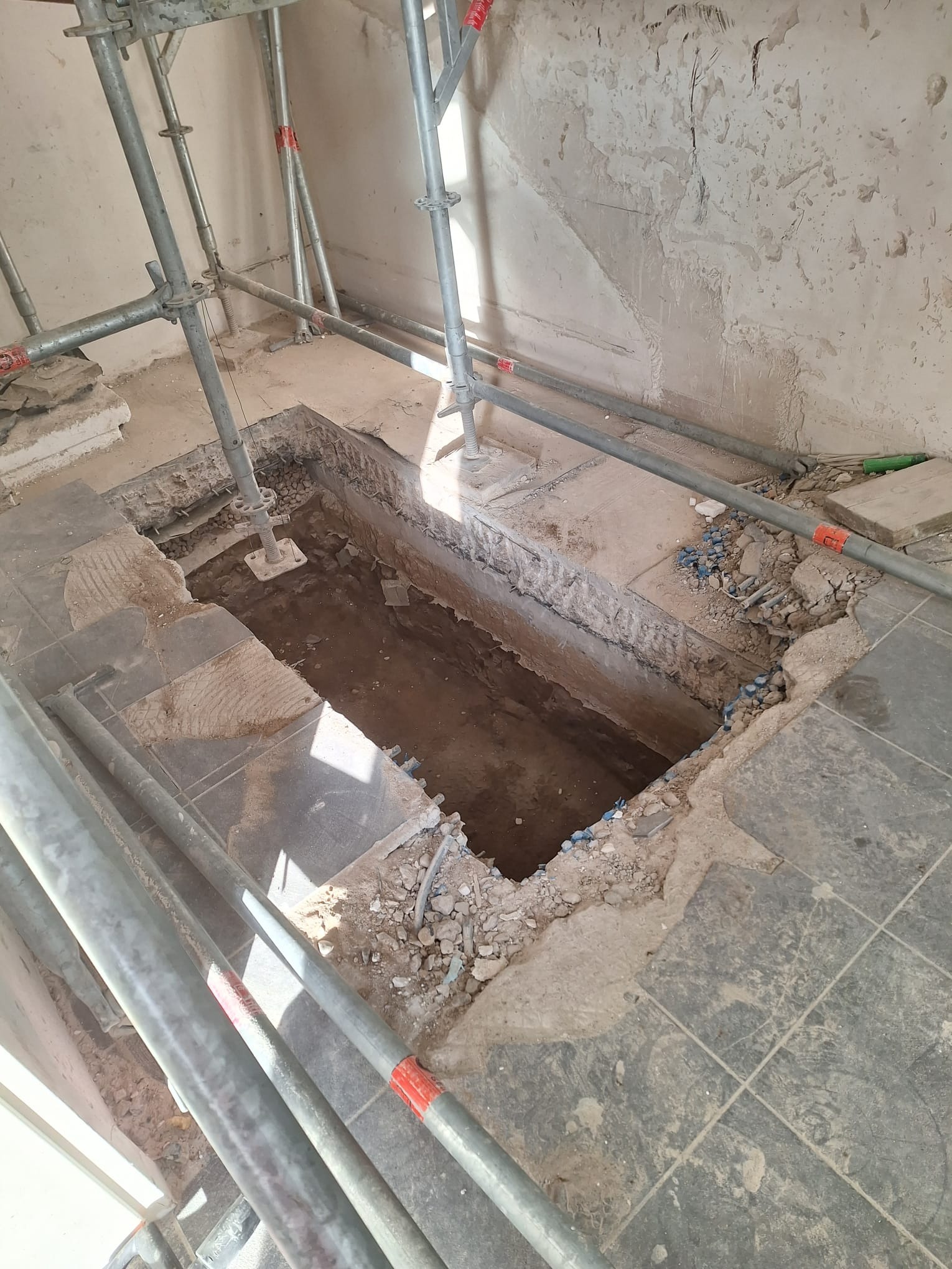 Wykop w posadzce z szarych płytek, widoczne fragmenty gruzu i instalacji, ściana z resztkami tynku, metalowe rusztowanie w pomieszczeniu w trakcie prac demontażowych.