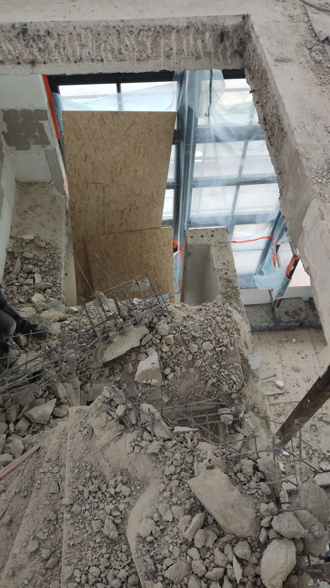 Gruz i odsłonięte zbrojenie pozostałe po częściowej rozbiórce ściany, widok z góry na usunięty fragment, z widoczną sklejką zabezpieczającą okno.