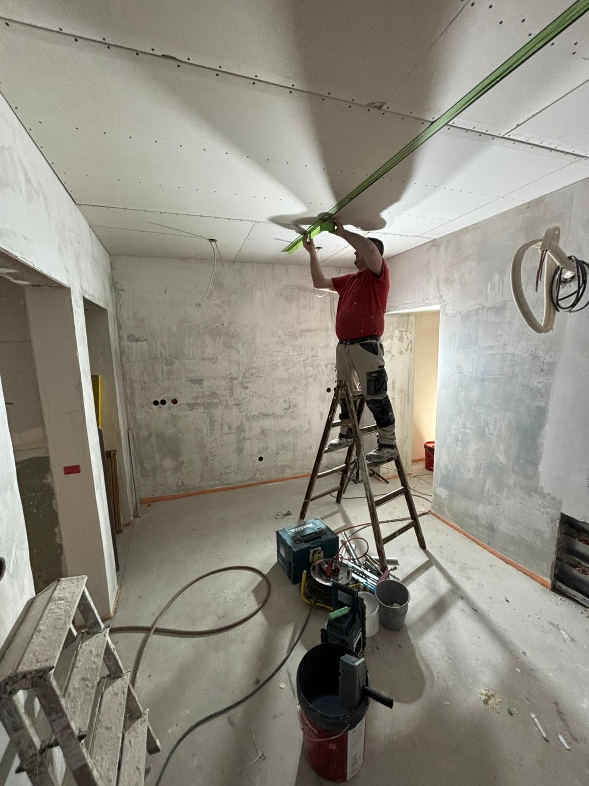 Pracownik na drabinie wyrównuje sufit z płyt gipsowo-kartonowych przy użyciu długiej, zielonej poziomicy w remontowanym pomieszczeniu z surowymi ścianami i widocznymi instalacjami.
