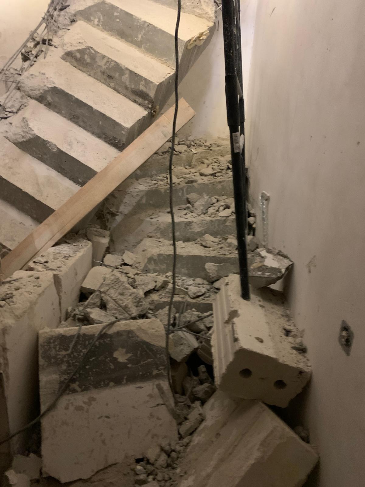Zdemontowane betonowe schody z widocznymi pozostałościami gruzu, drewnianą deską i czarnymi rurami przy ścianie.