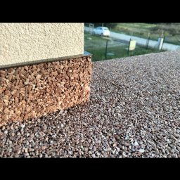 PK Biznes - Zewnętrzna ściana budynku z dekoracyjnym wykończeniem z drobnych kamyczków w odcieniach brązu, płynnie przechodząca w nawierzchnię z tego samego materiału, z widokiem na podjazd i zaparkowany...