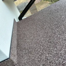 Wykończona powierzchnia balkonu z brązowego kruszywa kwarcowego, widoczna rynna odprowadzająca wodę i fragment elewacji budynku w tle.