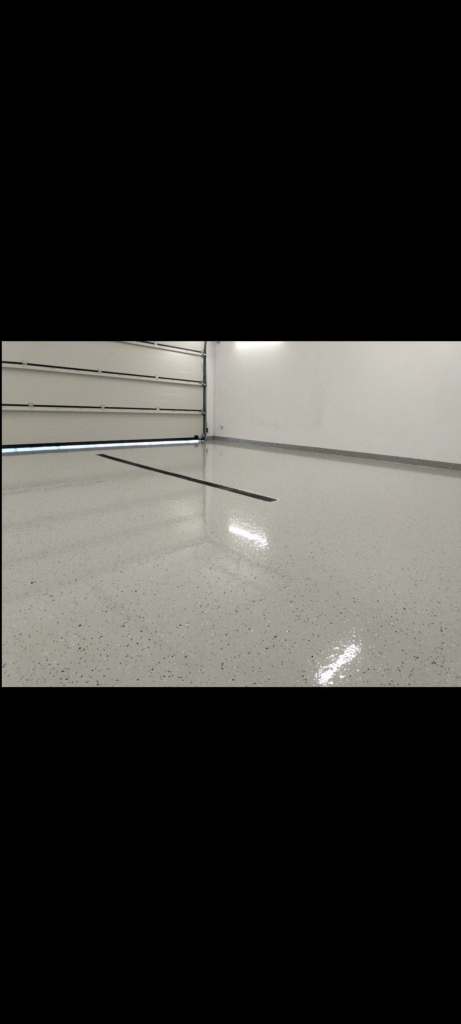 Jasnoszara, błyszcząca posadzka w garażu z widoczną szczeliną dylatacyjną i częściowo otwartą bramą garażową.