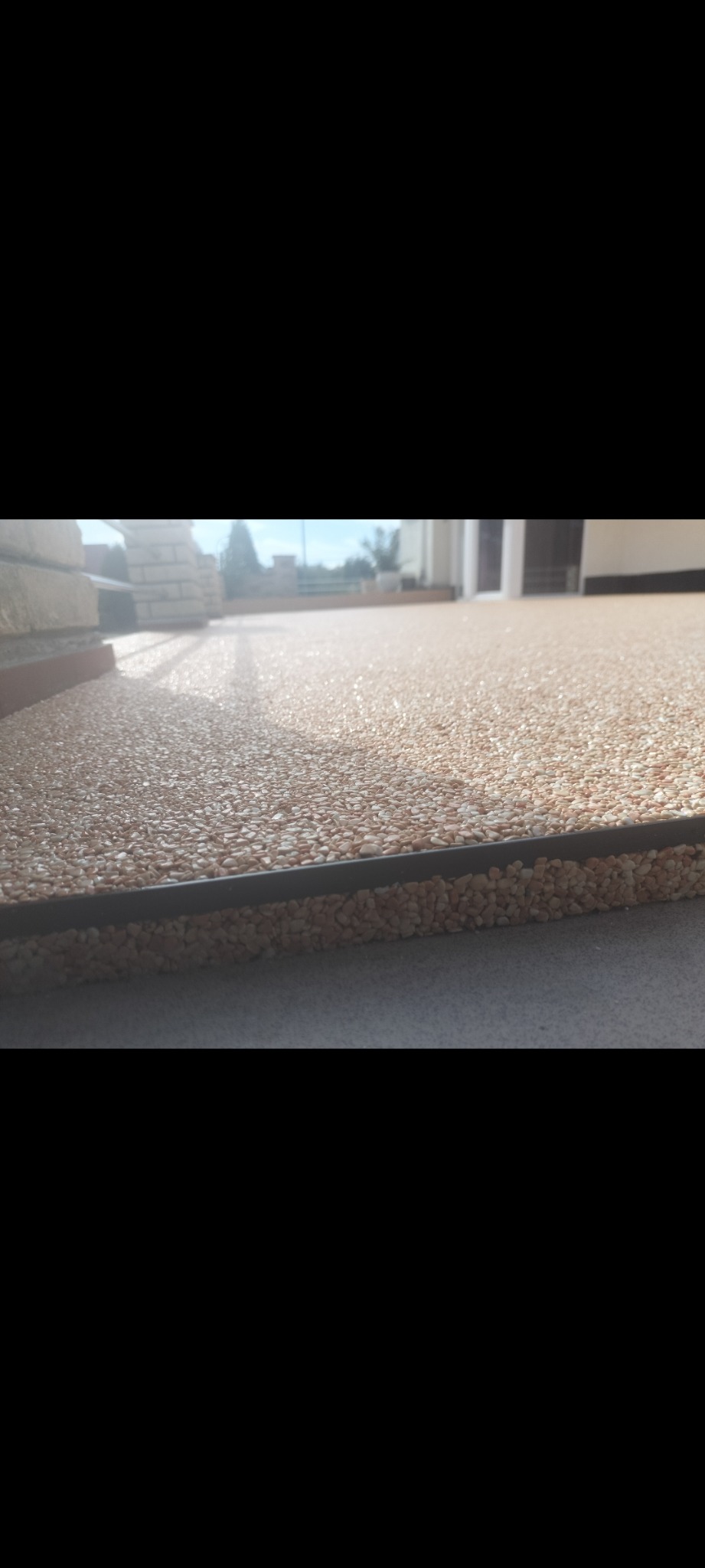 Beżowa posadzka z kamyczków na tarasie, widok z niskiej perspektywy, z widoczną listwą krawędziową i fragmentem elewacji budynku w tle.