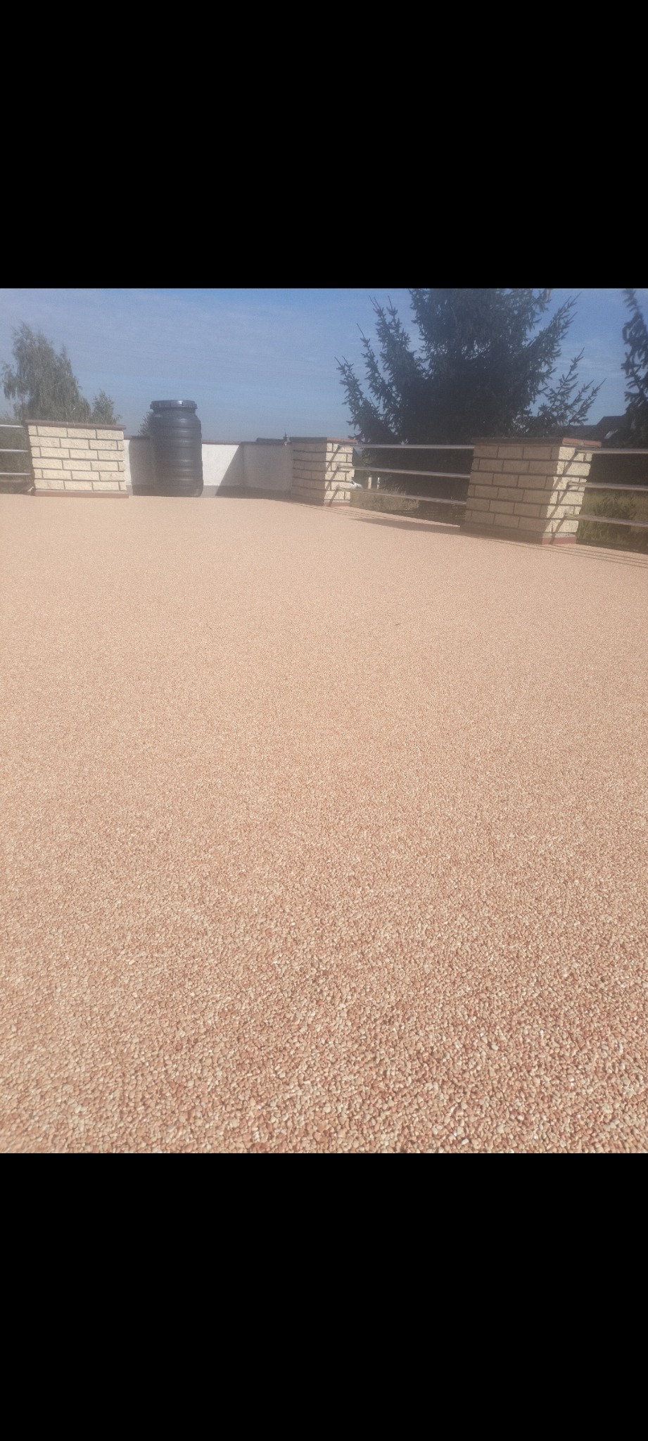 Beżowa, drobnoziarnista powierzchnia tarasu z niską ceglaną balustradą i metalową barierką, w tle zbiornik na wodę i zieleń.