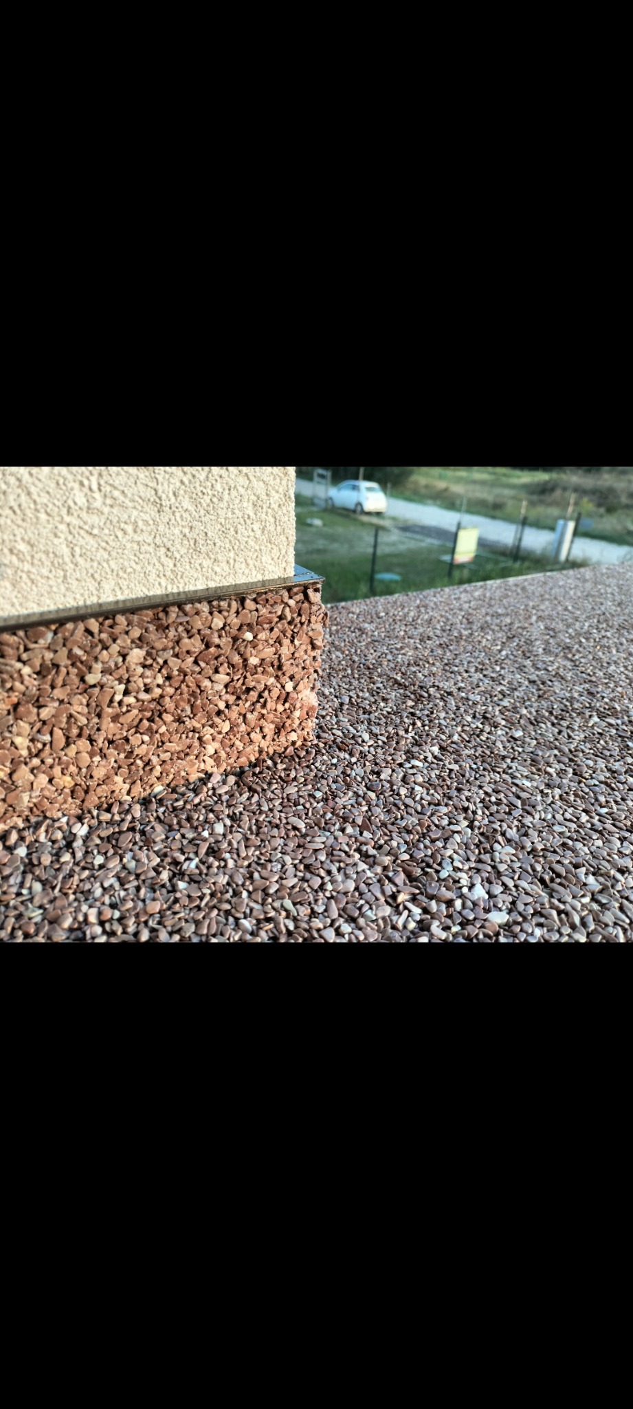 Zewnętrzna ściana budynku z dekoracyjnym wykończeniem z drobnych kamyczków w odcieniach brązu, płynnie przechodząca w nawierzchnię z tego samego materiału, z widokiem na podjazd i zaparkowany...