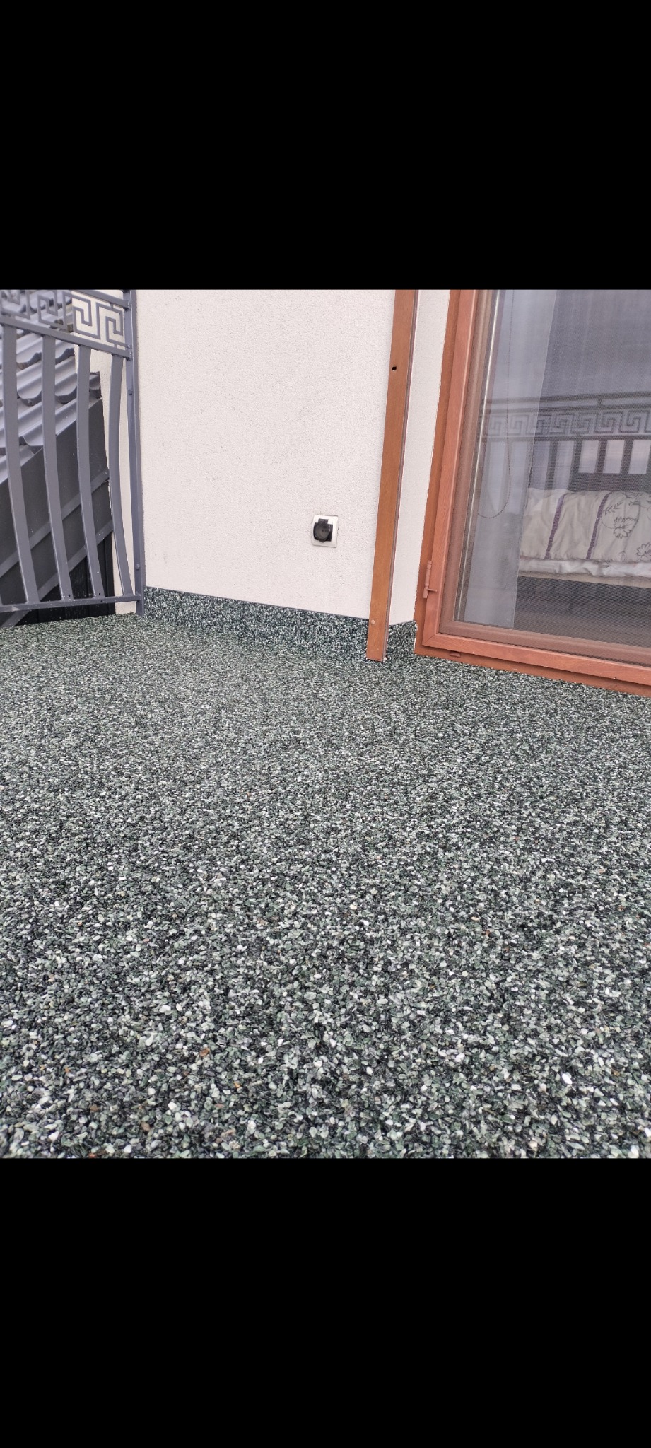 Szara posadzka z drobnych kamyczków na balkonie, widoczna ściana z gniazdkiem elektrycznym i fragment balustrady.