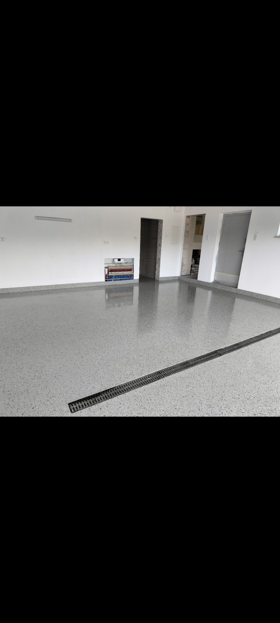 Szara, błyszcząca posadzka z odpływem liniowym w nowo wykończonym pomieszczeniu z białymi ścianami i instalacją centralnego ogrzewania.