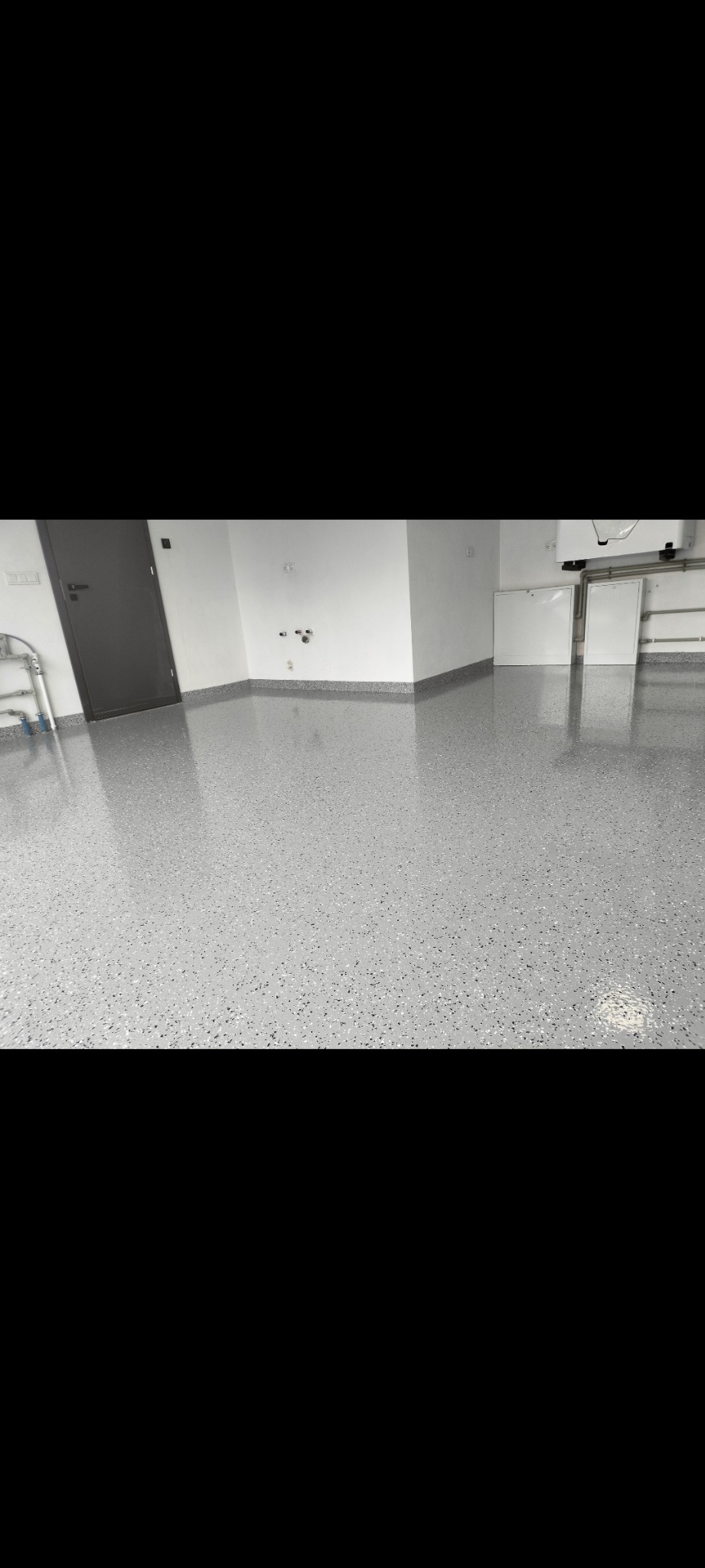 Jasnoszara, błyszcząca podłoga z drobnymi, ciemnymi plamkami w nowo wykończonym garażu z białymi ścianami i szarą, metalową bramą w tle.