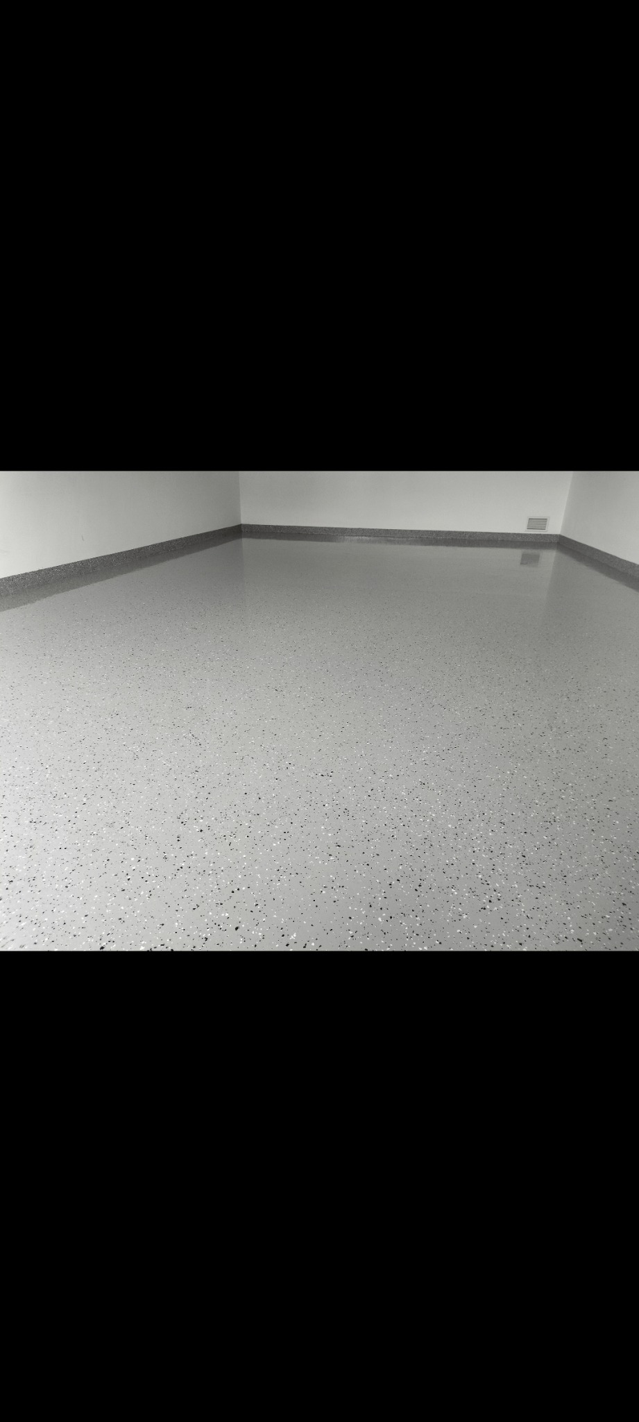 Jasnoszara posadzka z drobnymi, czarnymi i białymi inkluzjami, wykonana w rogu pomieszczenia, widoczna kratka wentylacyjna przy podłodze.