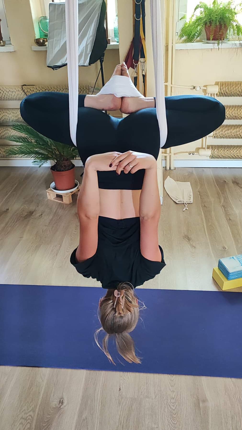 Kobieta w czarnym stroju wykonuje pozycję odwróconą w hamaku do aerial jogi, stopy zabezpieczone materiałem, na podłodze fioletowa mata do ćwiczeń.