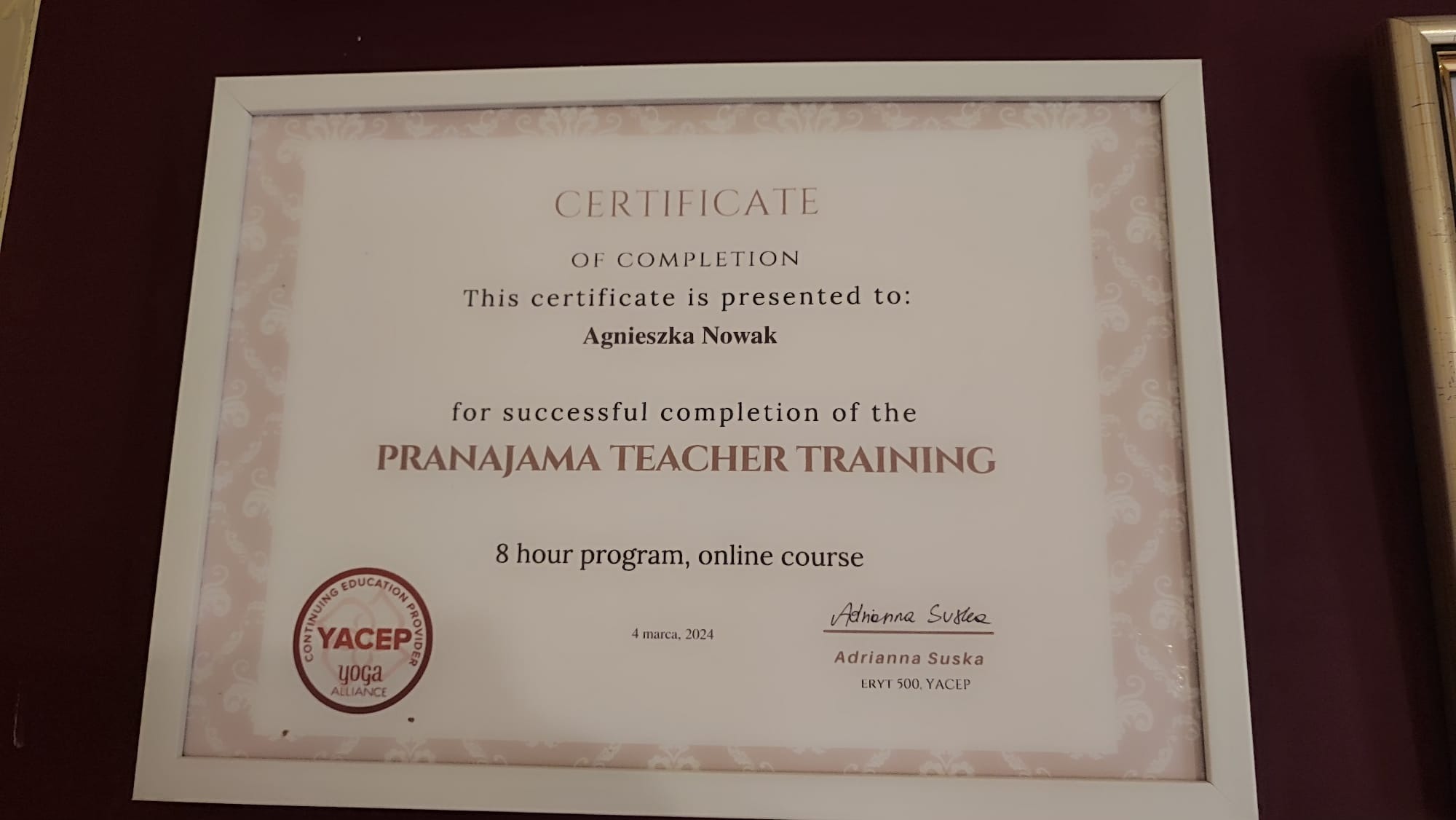 Certyfikat ukończenia kursu Pranjajama Teacher Training dla Agnieszki Nowak, oprawiony w białą ramkę, wiszący na ścianie o bordowym kolorze.