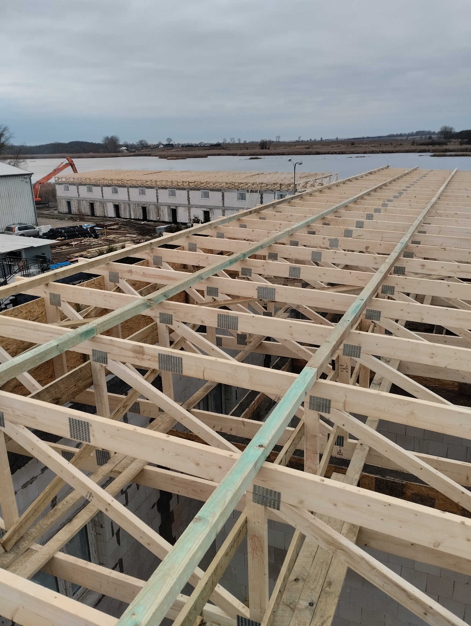 Widok z góry na konstrukcję drewnianą dachu w trakcie budowy, z widocznymi elementami więźby dachowej i metalowymi złączami, w tle budynek w stanie surowym otwartym oraz krajobraz z jeziorem...