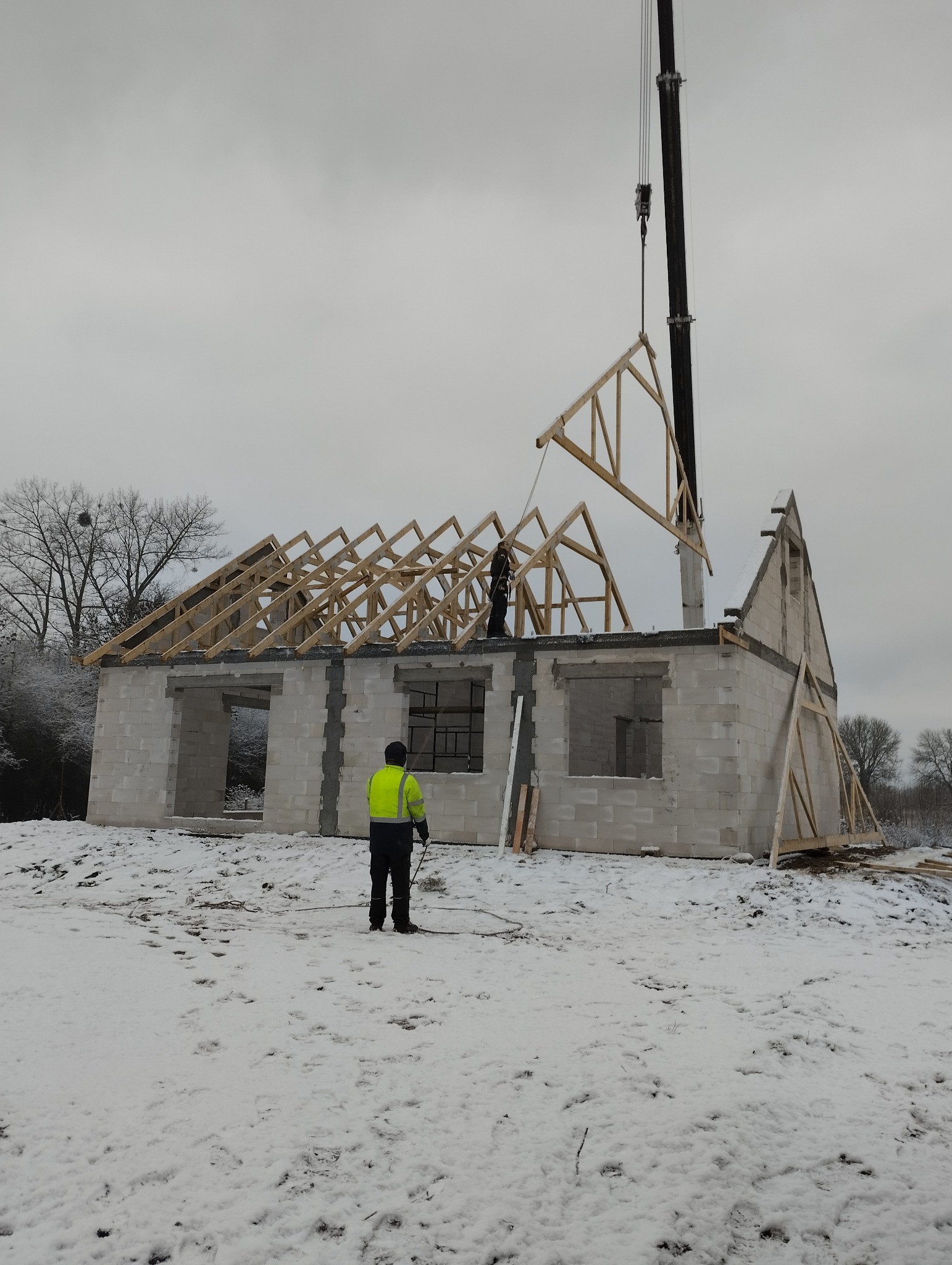 Montaż drewnianej konstrukcji dachu na surowym budynku z bloczków, widoczny dźwig i pracownicy w zimowej scenerii.