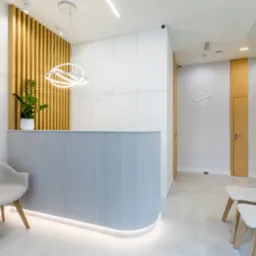 Jasne, nowoczesne wnętrze recepcji z szarym biurkiem, drewnianymi lamelami, okrągłą lampą i minimalistycznymi krzesłami w poczekalni, logo gabinetu na ścianie.