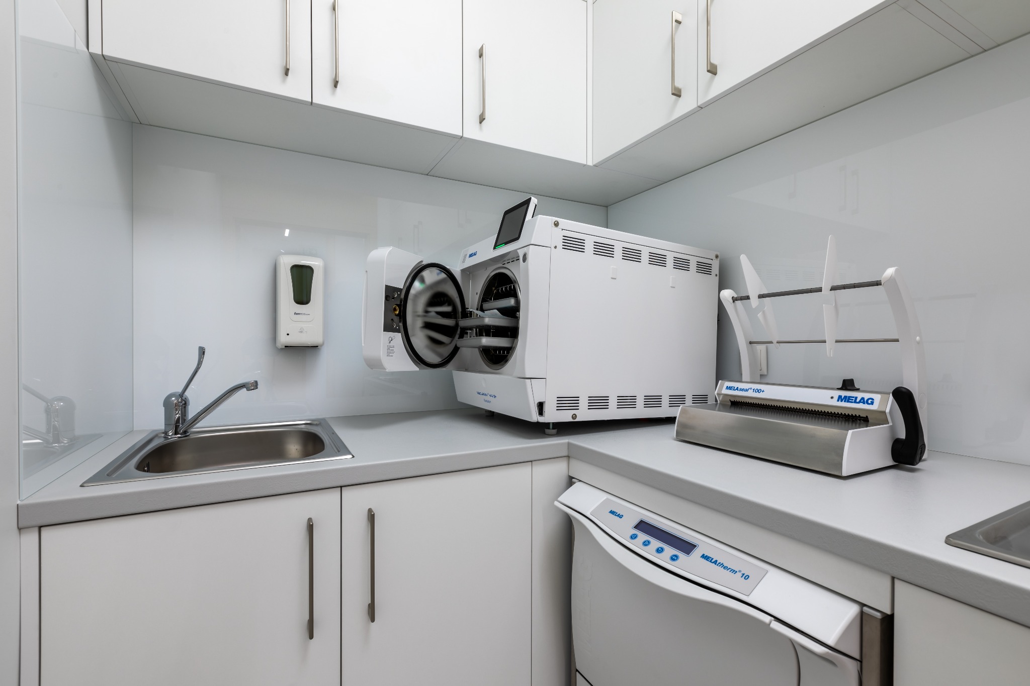 Sterylne, białe wnętrze gabinetu z autoklawem Melag z otwartymi drzwiami, zlewem, dozownikiem mydła i zgrzewarką do pakietów.
