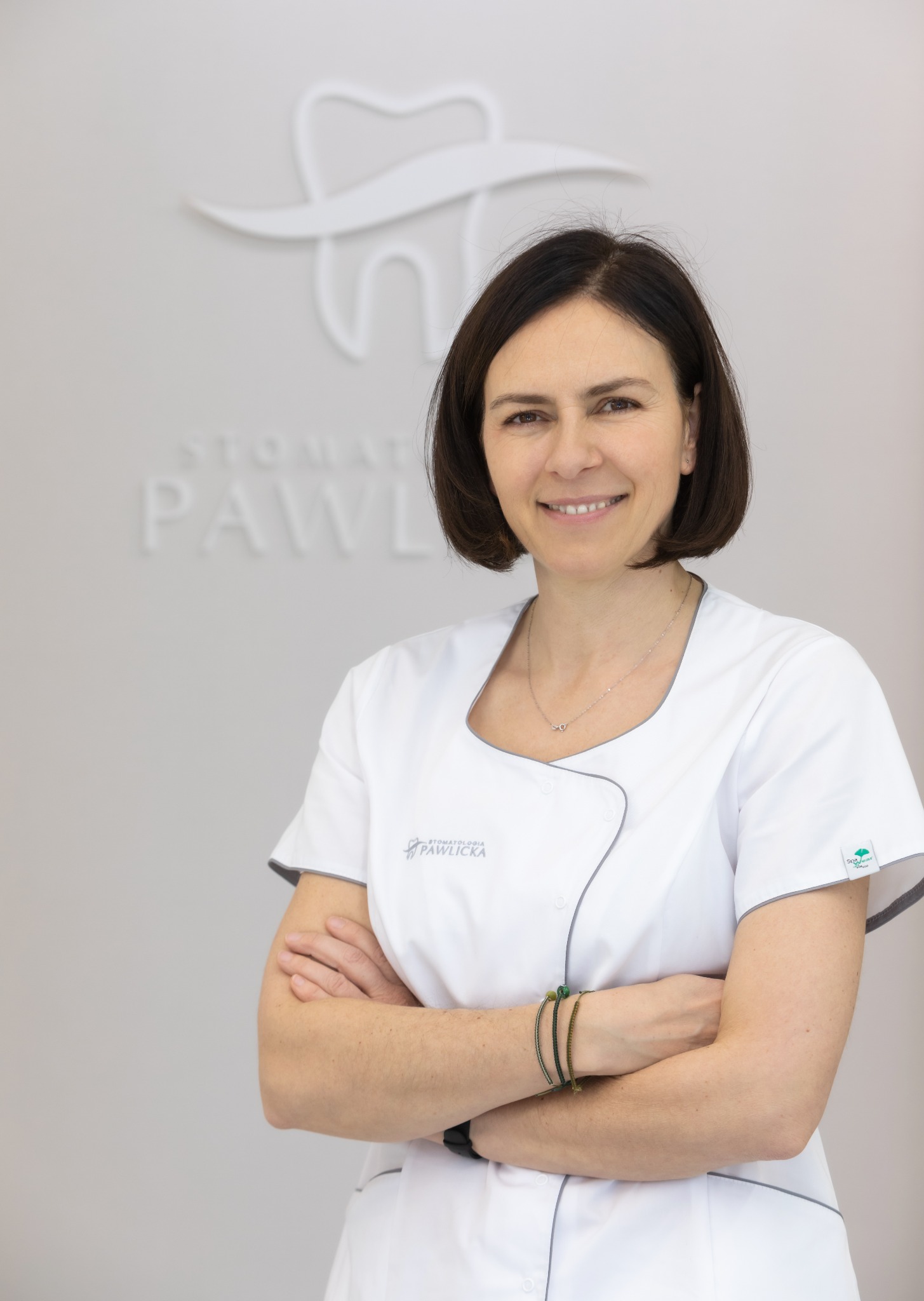 Uśmiechnięta kobieta w białym uniformie z logo kliniki na tle minimalistycznego wnętrza gabinetu stomatologicznego z logo zęba.